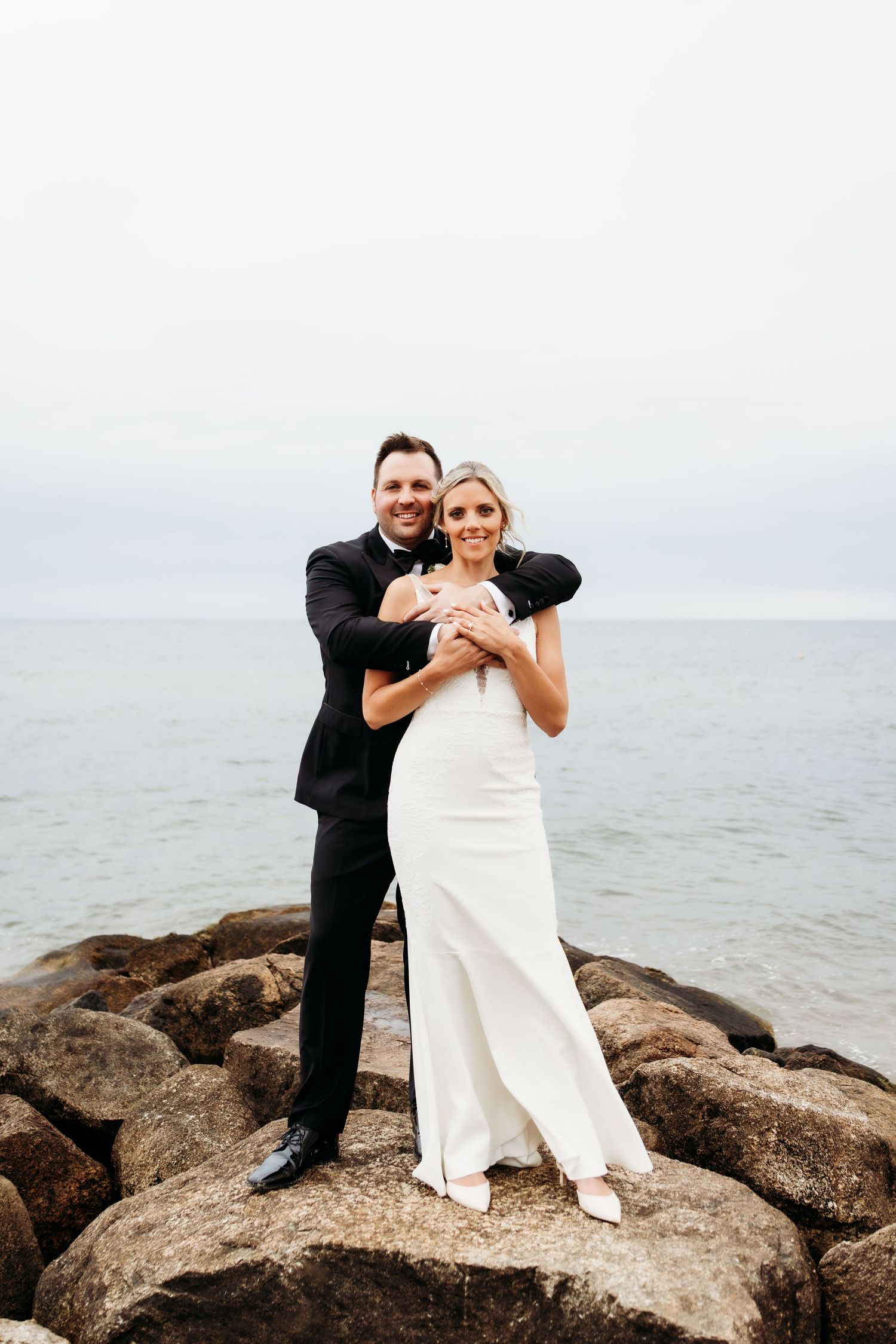 A couple in wedding attire shares a playful moment while posing on coastal rocks by the ocean.