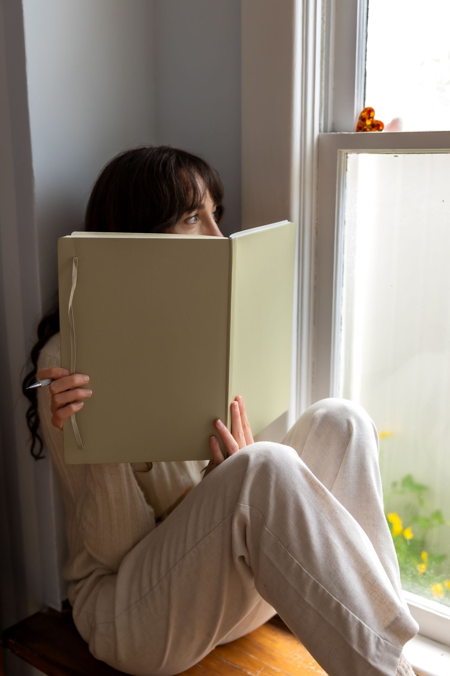 Someone in a white sweater reads a book by a bright window with yellow flowers visible outside.
