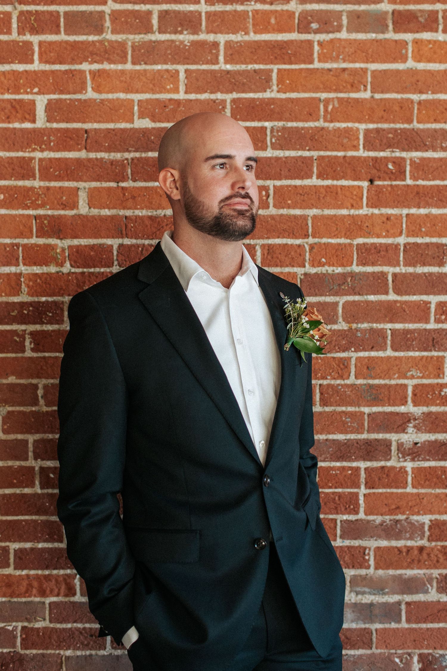 Classic groom's attire featuring a black tuxedo and white boutonniere posed against rustic brick walls.