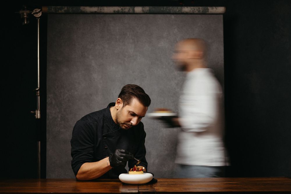 A person in dark clothing smells a bowl of food in a dimly lit setting with dramatic lighting.