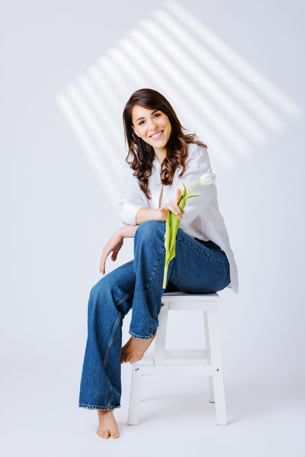Someone in casual denim and white shirt sits barefoot on white chair holding single tulip in minimalist setting.