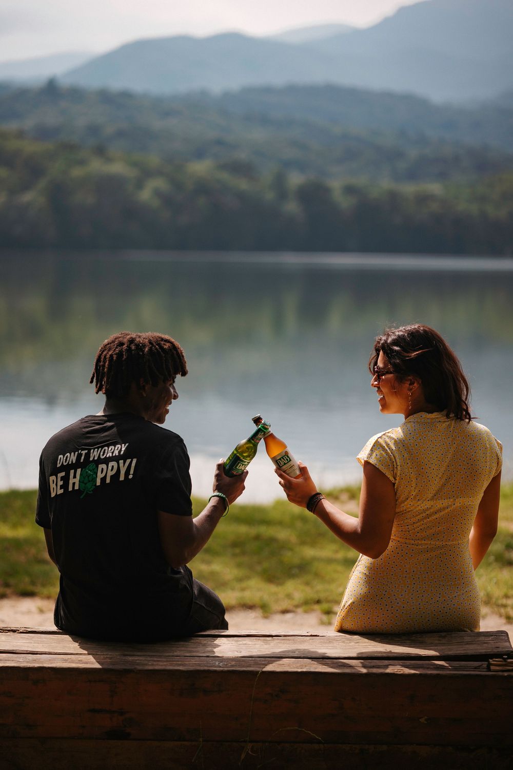 Two friends sit side by side on a wooden dock overlooking a serene lake surrounded by mountains in the background.