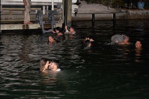 A group of people swim and splash in dark waters near a wooden dock during a summer evening.