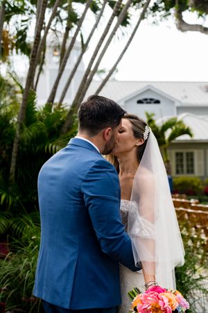 Couple shares a romantic kiss in a lush garden setting with a white building in the background.