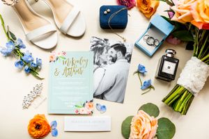 Wedding details flatlay with blue and peach flowers, invitations, and accessories on white background.
