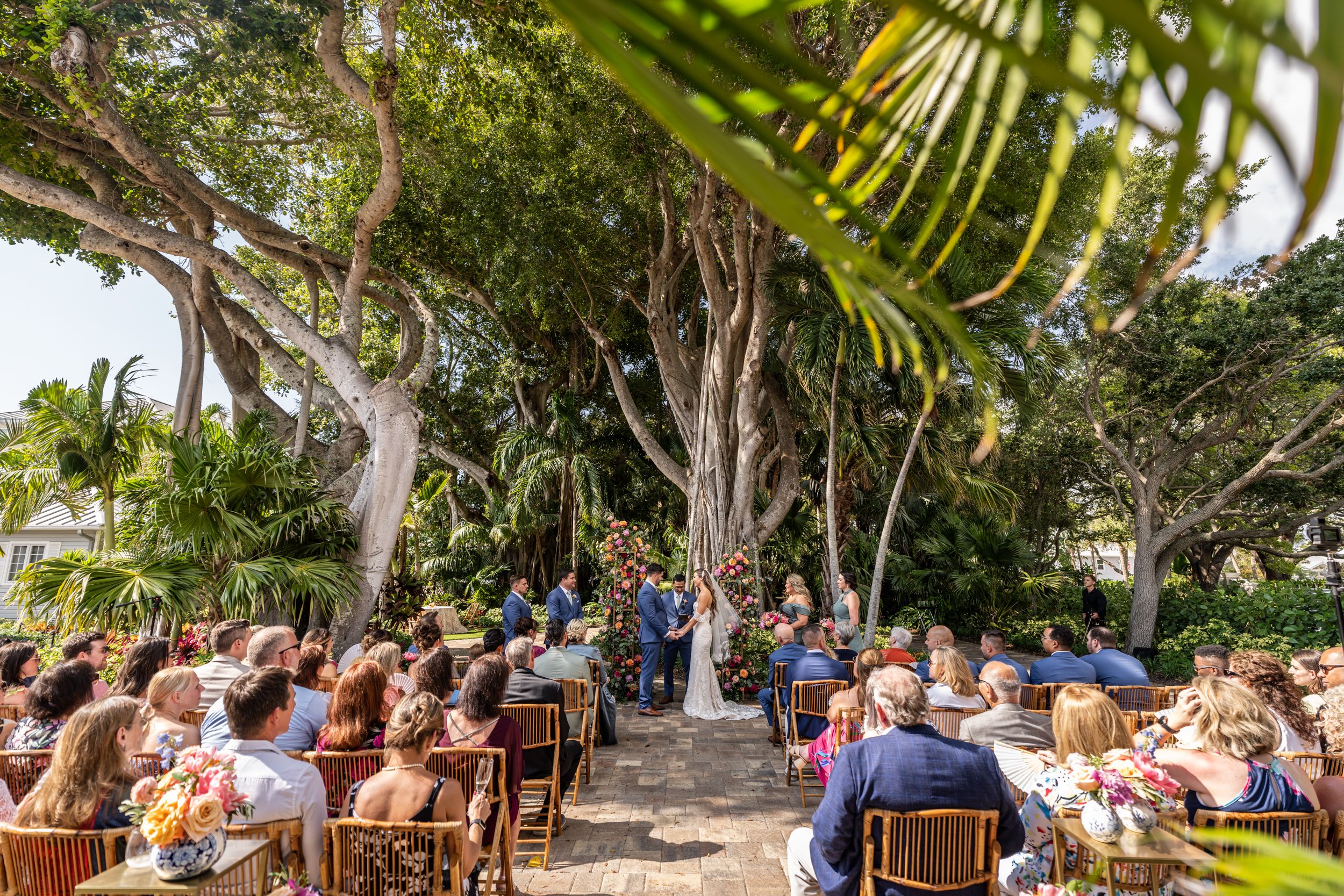 Wedding ceremony setup in a tropical garden setting with rows of white chairs under palm trees.
