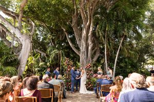Outdoor wedding ceremony setup with wooden chairs under large trees.