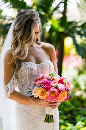 Close-up details of bride holding vibrant pink and orange rose wedding bouquet against lace dress.