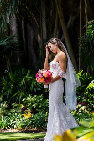 Bride in lace dress holding colorful bouquet in a tropical garden setting.