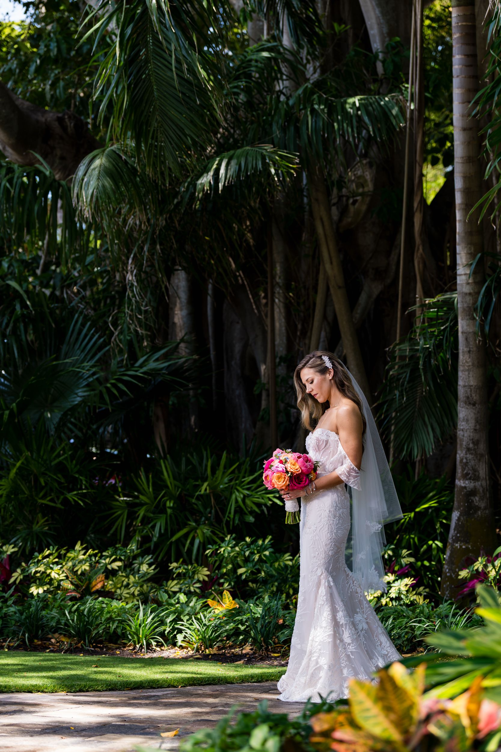 A bride in a lace wedding dress holds a pink bouquet while standing in a tropical garden setting.
