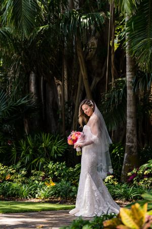 A white wedding dress flows elegantly against a lush tropical garden backdrop with palm trees.