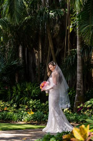 Bride in lace wedding dress poses in a lush tropical garden setting with palm trees and colorful flowers.
