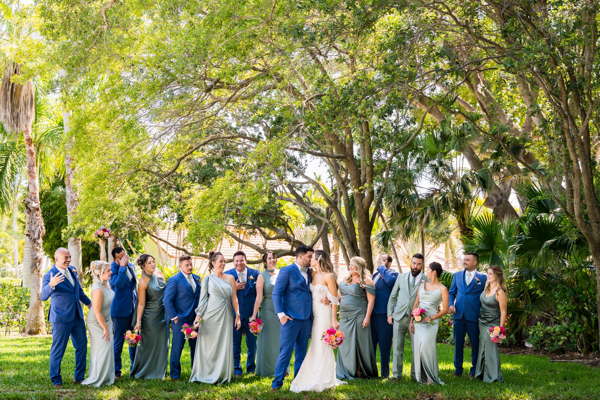 Wedding party in navy blue and gray poses under a canopy of green trees.