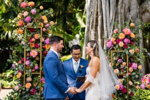 Wedding ceremony under floral arches with vibrant pink and orange roses in a lush garden setting.