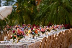 Long wedding reception table set under palm trees with colorful floral centerpieces and wooden chairs.