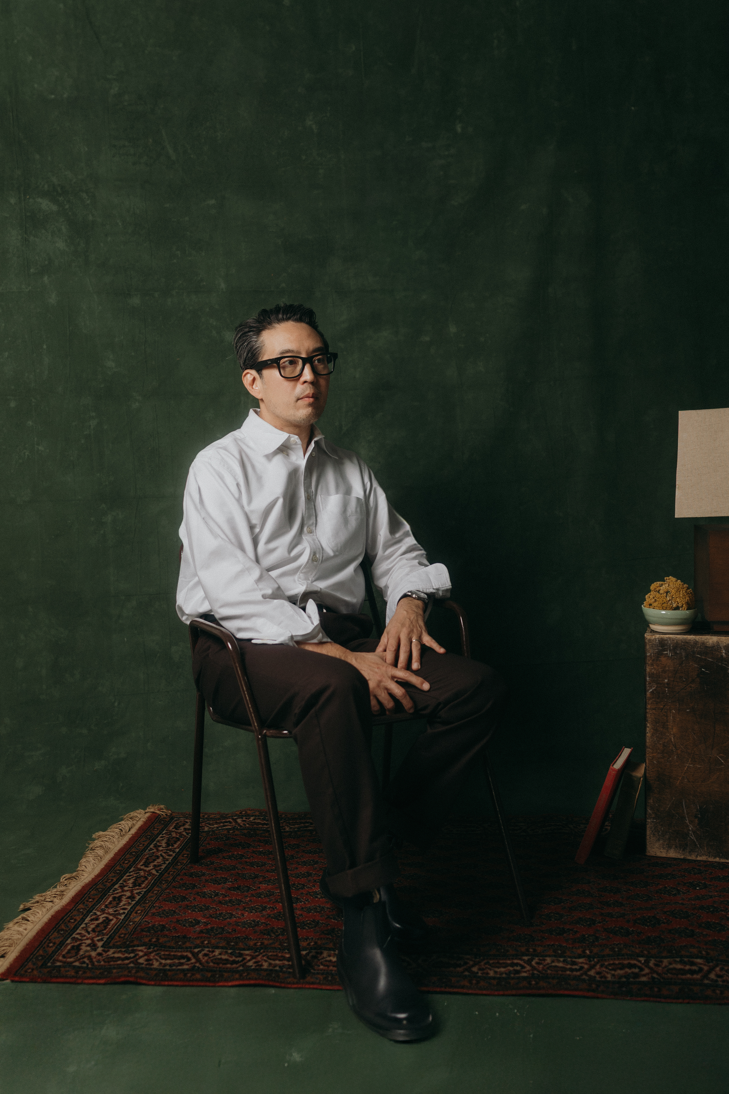 A person in a white dress shirt and tie sits on a chair against a dark green wall with an oriental rug beneath.