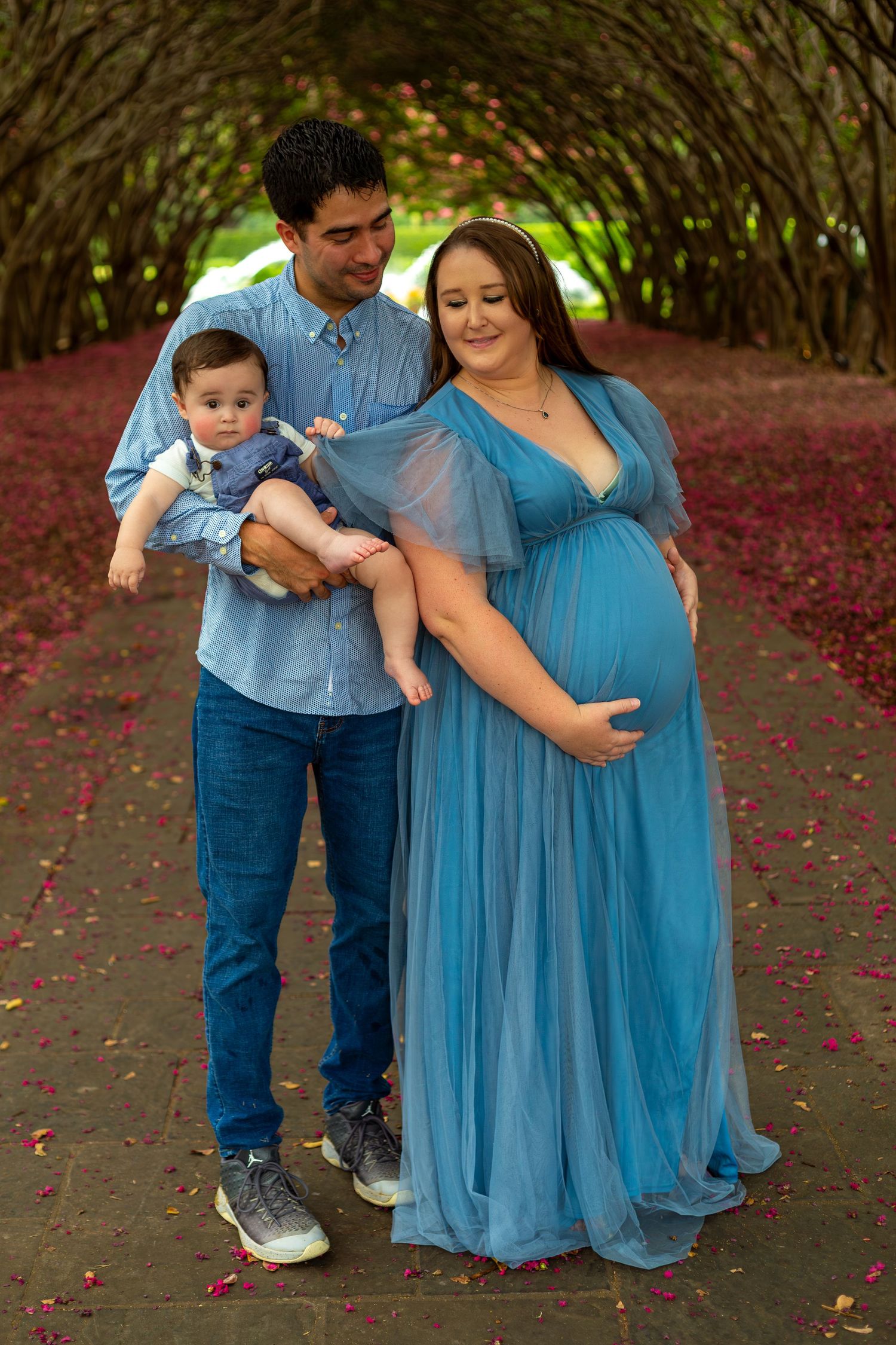 A family poses under a garden tunnel archway with red petals scattered on the ground, wearing coordinating blue outfits.