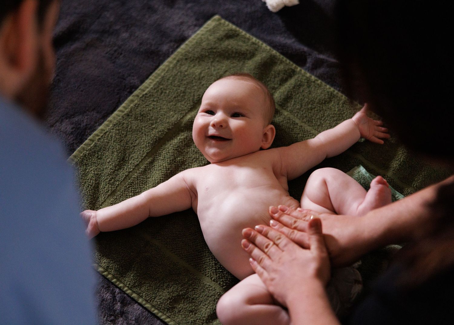 Baby lying on dark green blanket while adult hands reach to tickle their tummy.