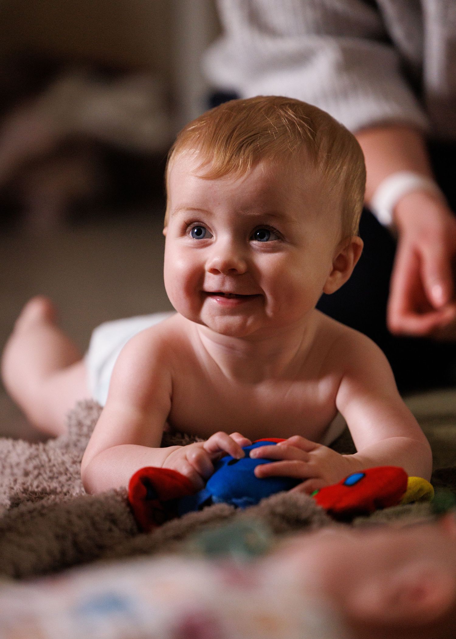 A baby lying on a soft surface plays with colorful toys while looking off to the side with a gentle smile.