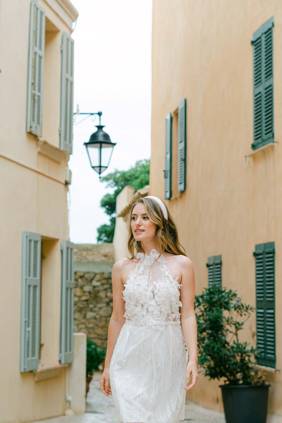 Mariée en robe blanche dans une ruelle provençale aux volets bleus, lumière douce, style fine art.