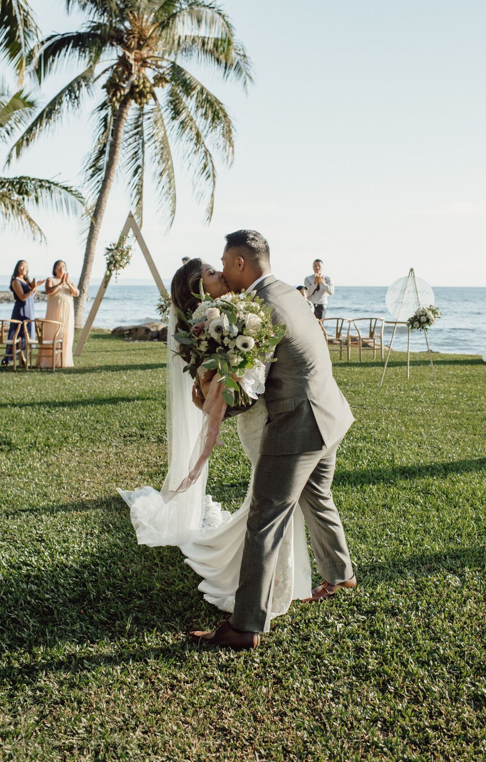 Couple dances on lush green lawn with palm trees and ocean in background.