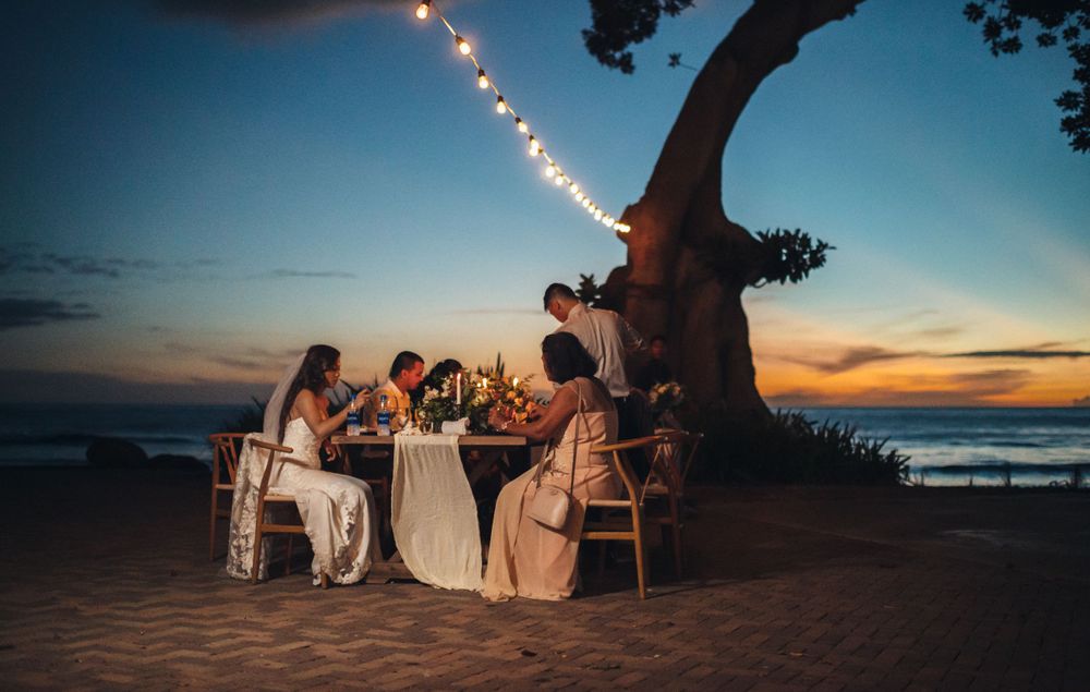 String lights illuminate a romantic beach wedding reception at sunset with tables set up on the sand.