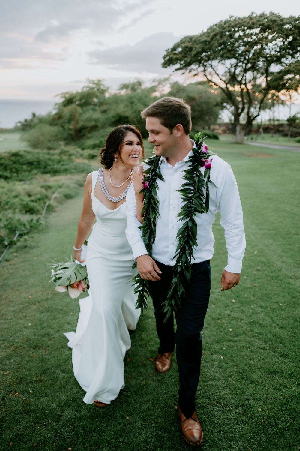 Newlyweds walk and laugh together on a grassy oceanside path.