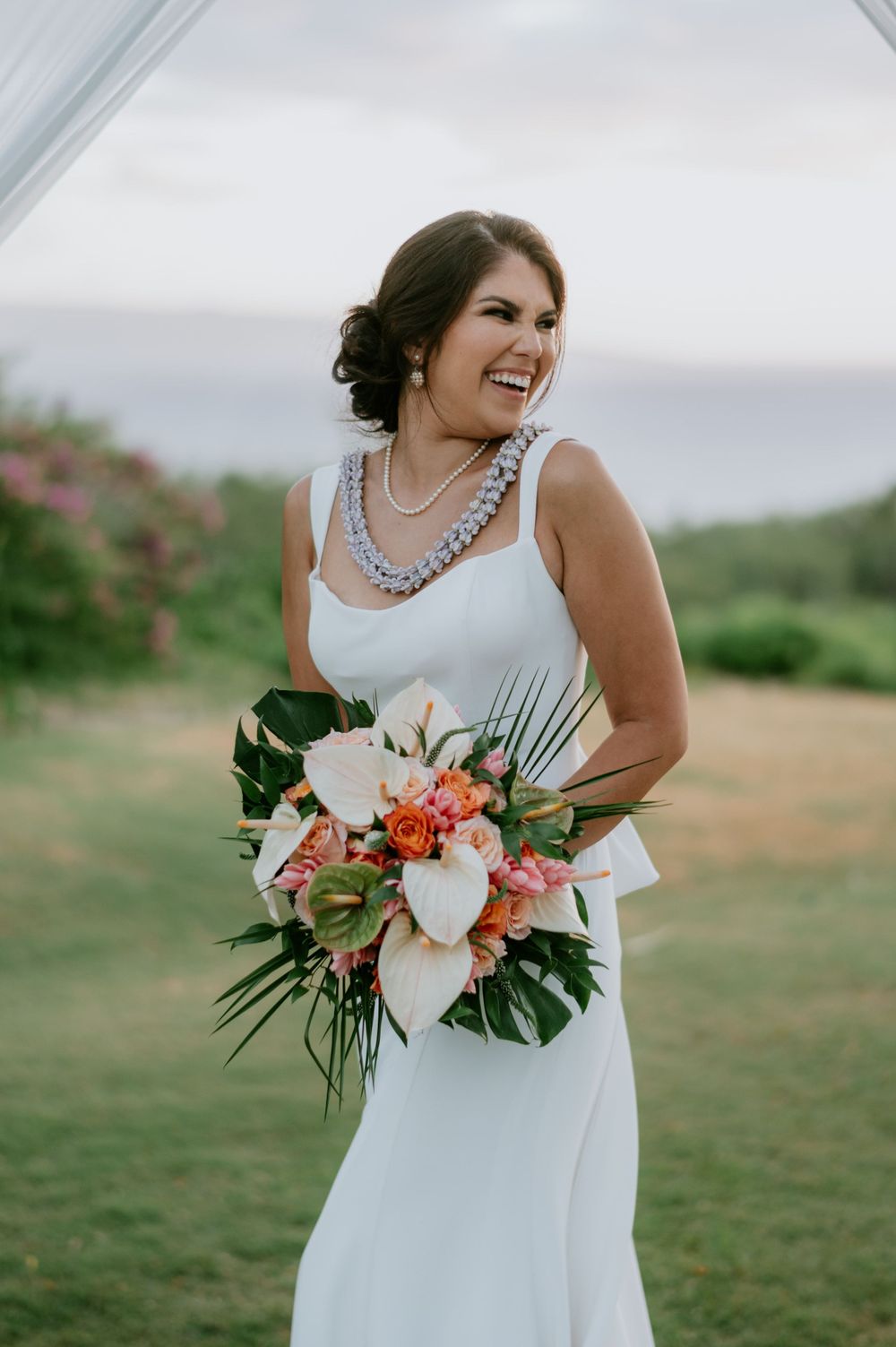 A beautiful bridal bouquet featuring coral and white flowers with tropical greenery against a grassy outdoor setting.