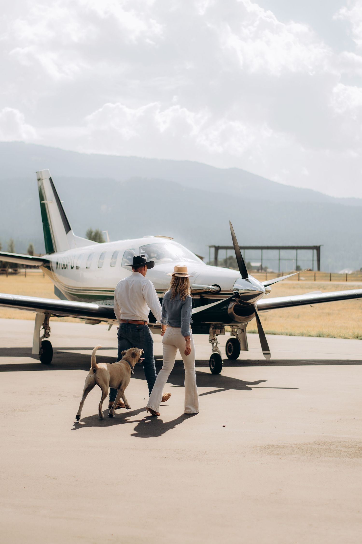 Two people walking towards a small private aircraft parked on an airfield with mountains visible in the background.