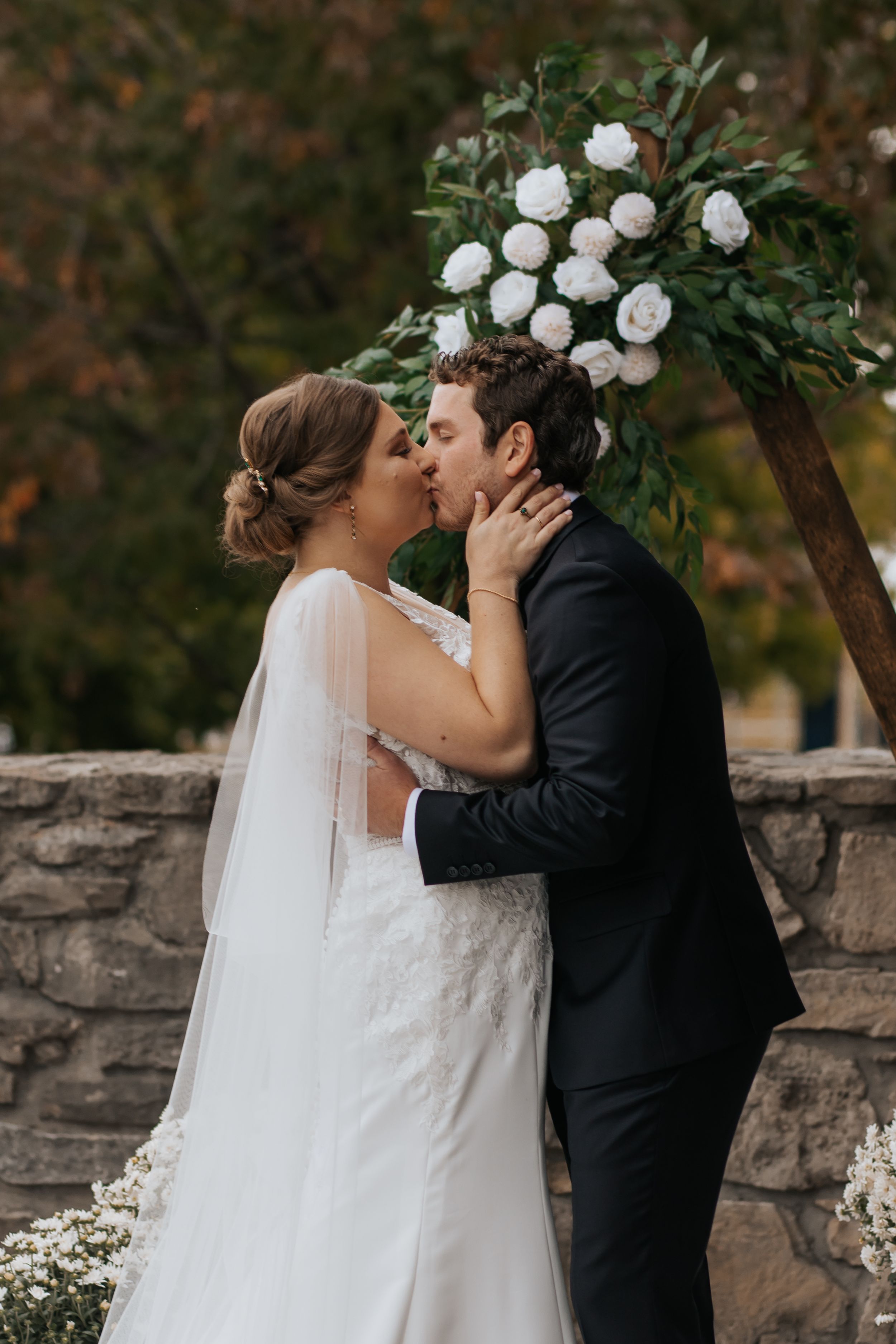 A romantic wedding couple shares a kiss in front of a stone wall decorated with white flowers and greenery.