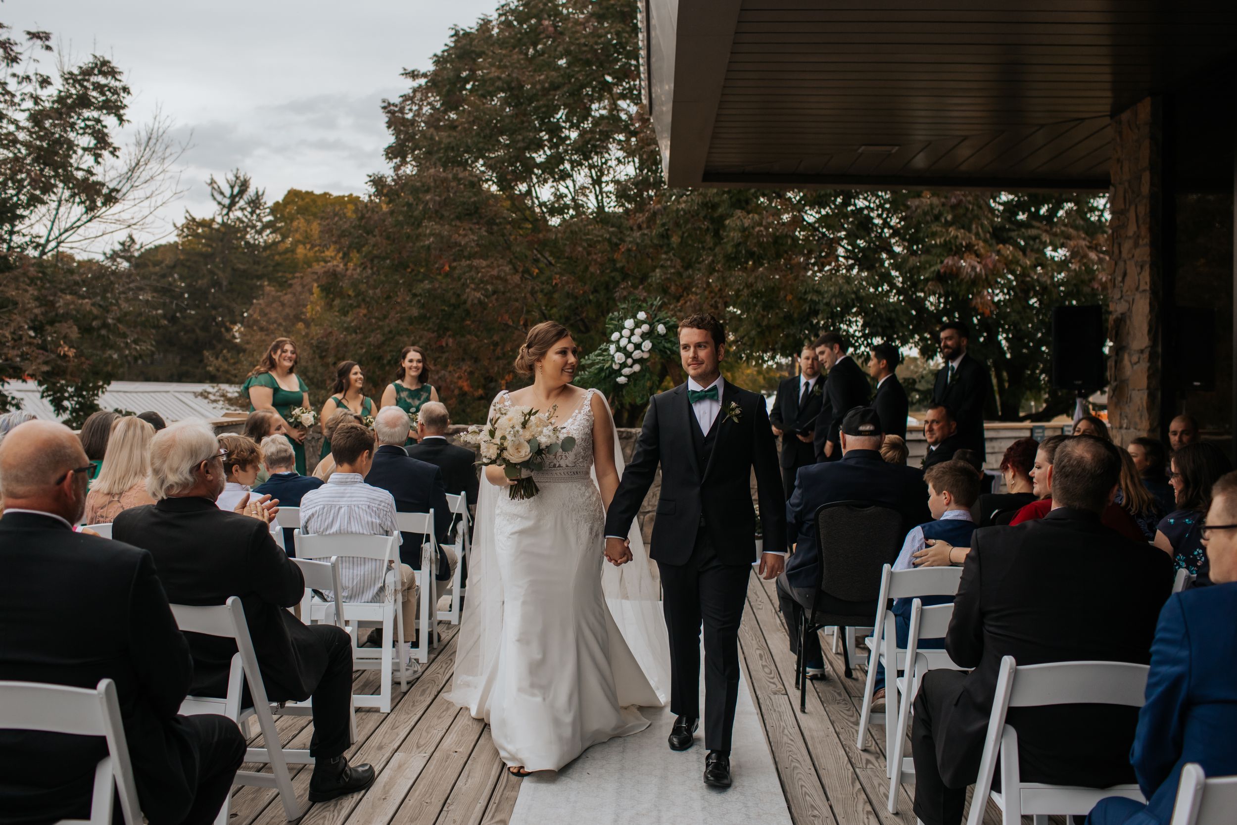Wedding ceremony guests seated in white chairs on an outdoor patio surrounded by autumn trees. Conner Prairie, Fishers Indiana