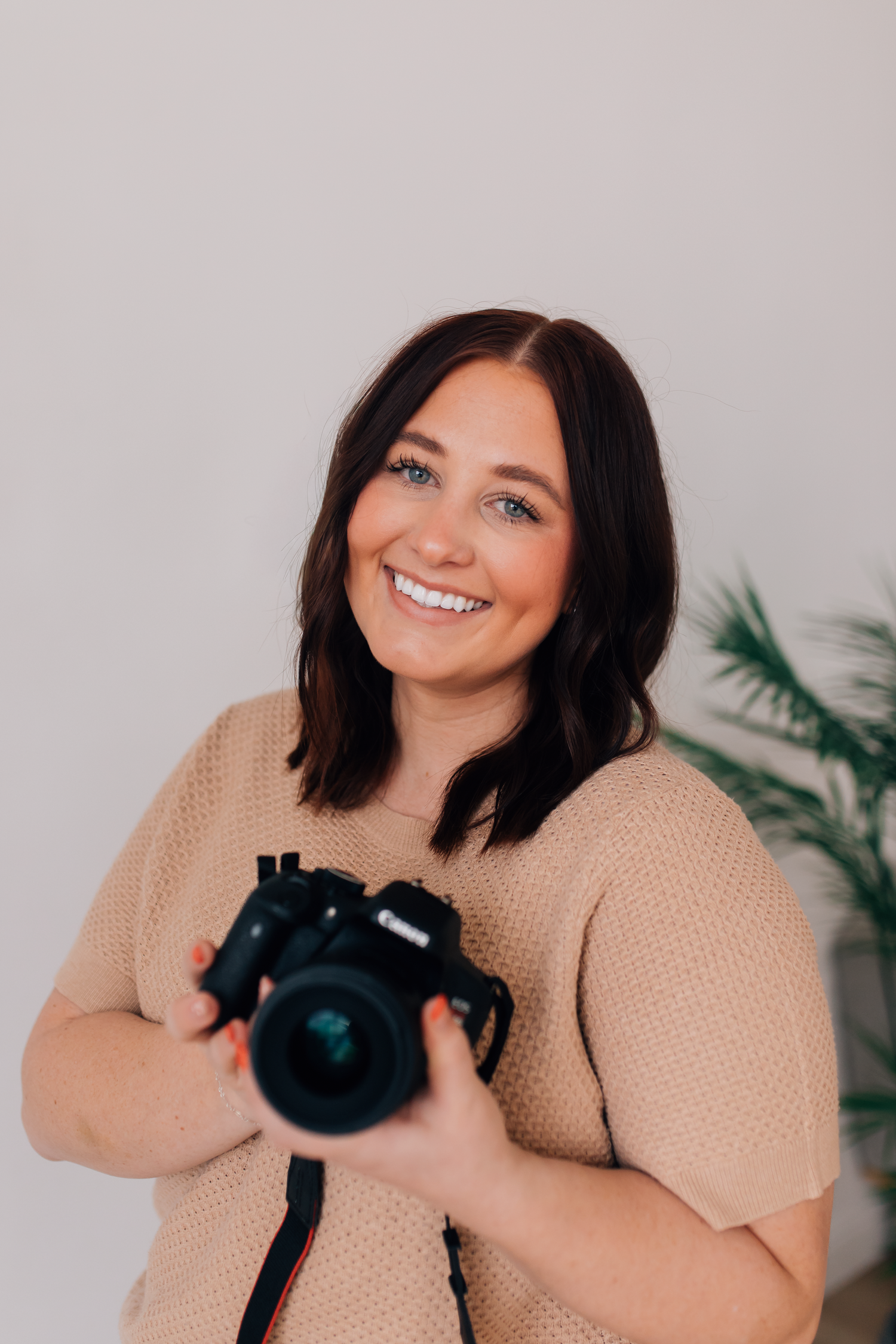 A person in a beige sweater smiles and poses with a camera in a bright studio setting with tropical plants.