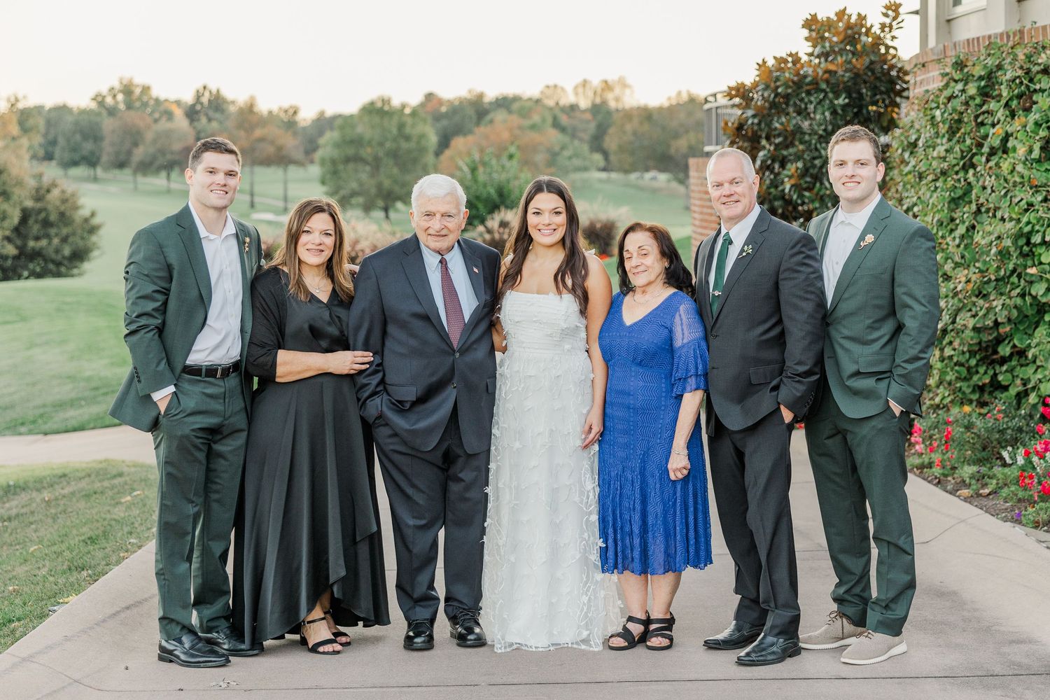 A group of wedding guests in formal attire stand together outdoors on a grassy hillside for a photo.