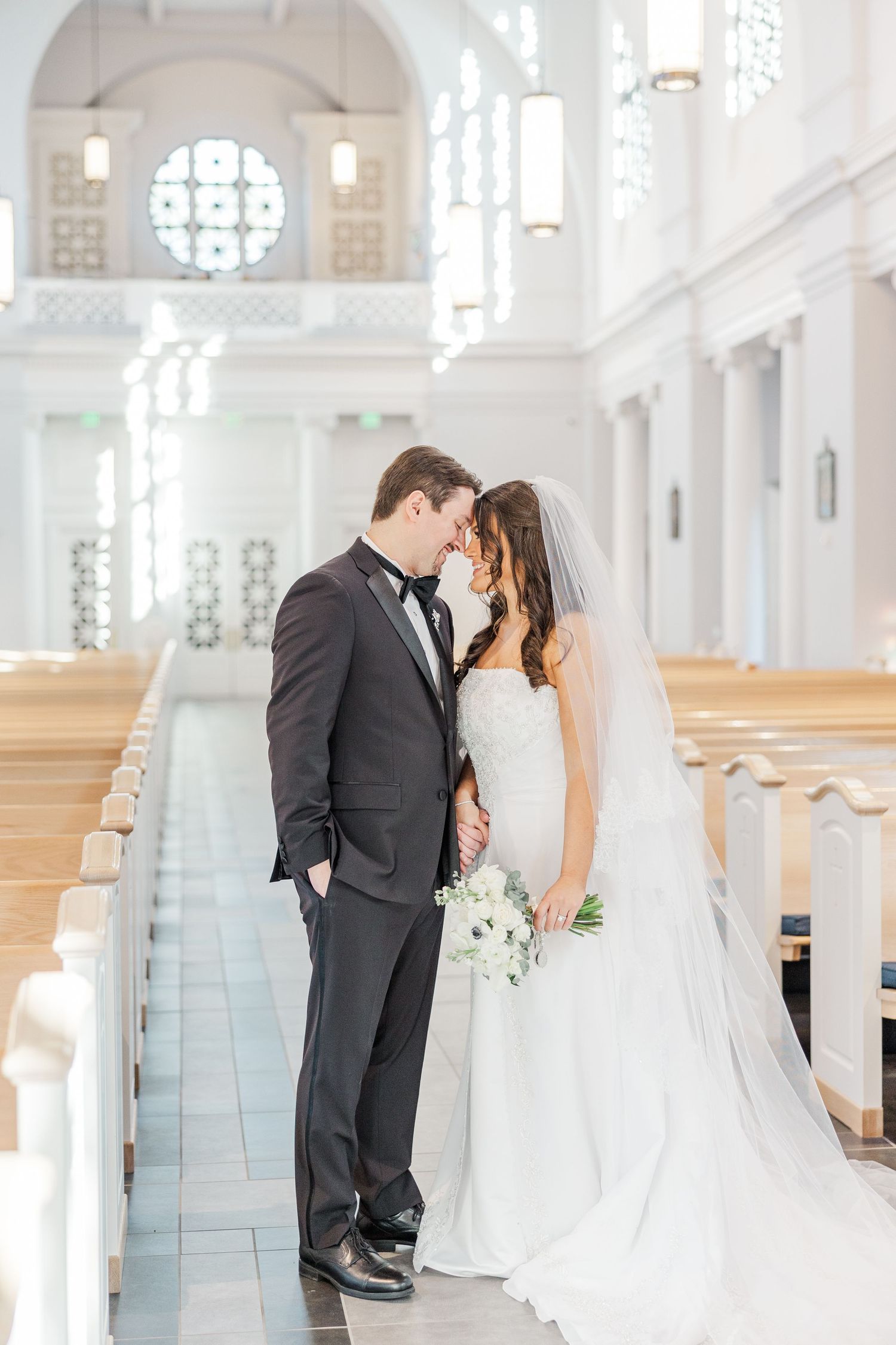 Romantic wedding couple shares intimate moments in bright white church interior with elegant columns and natural light.