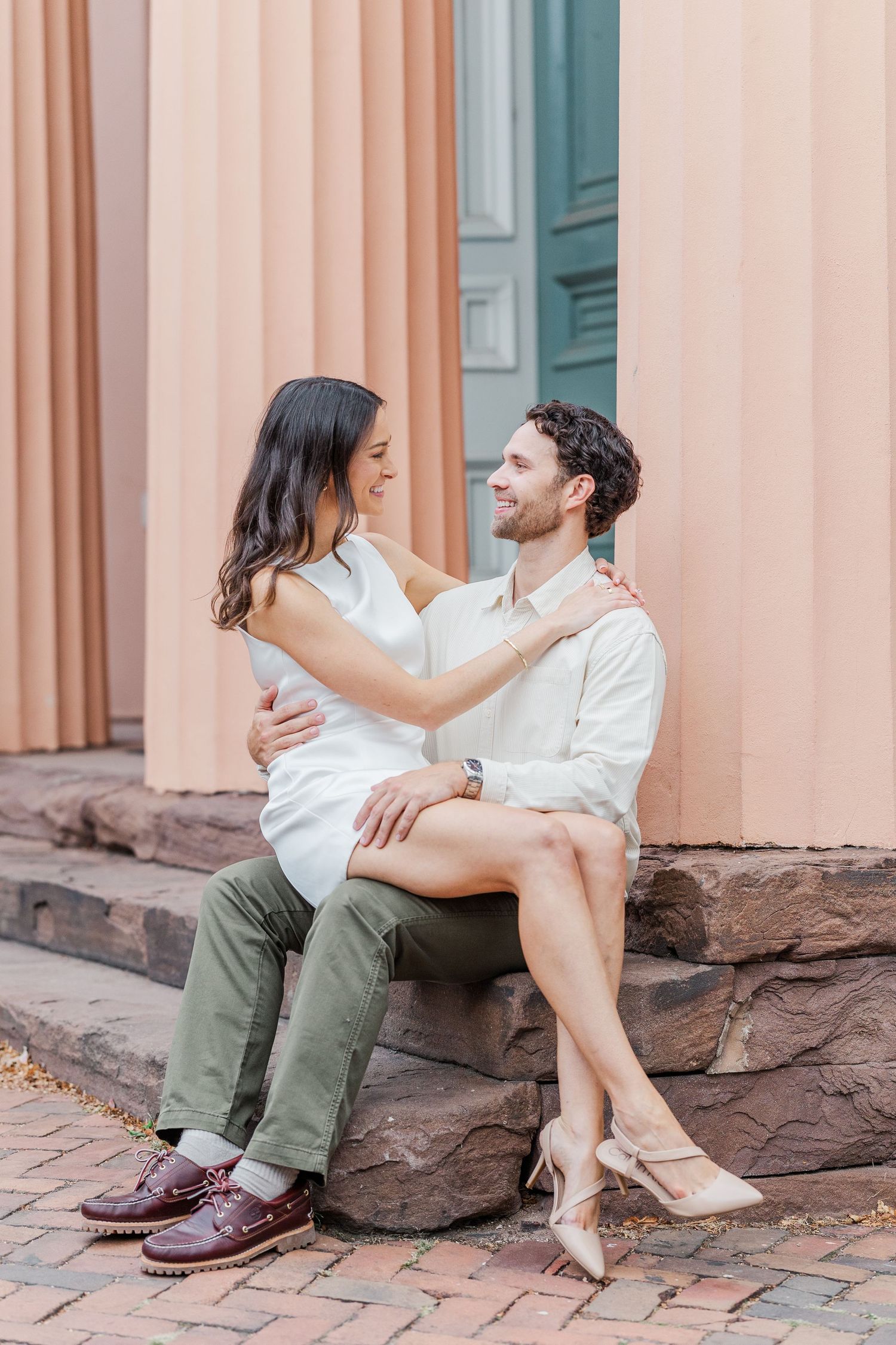 A couple sits together on stone steps wearing white tops and olive pants while sharing intimate moments.