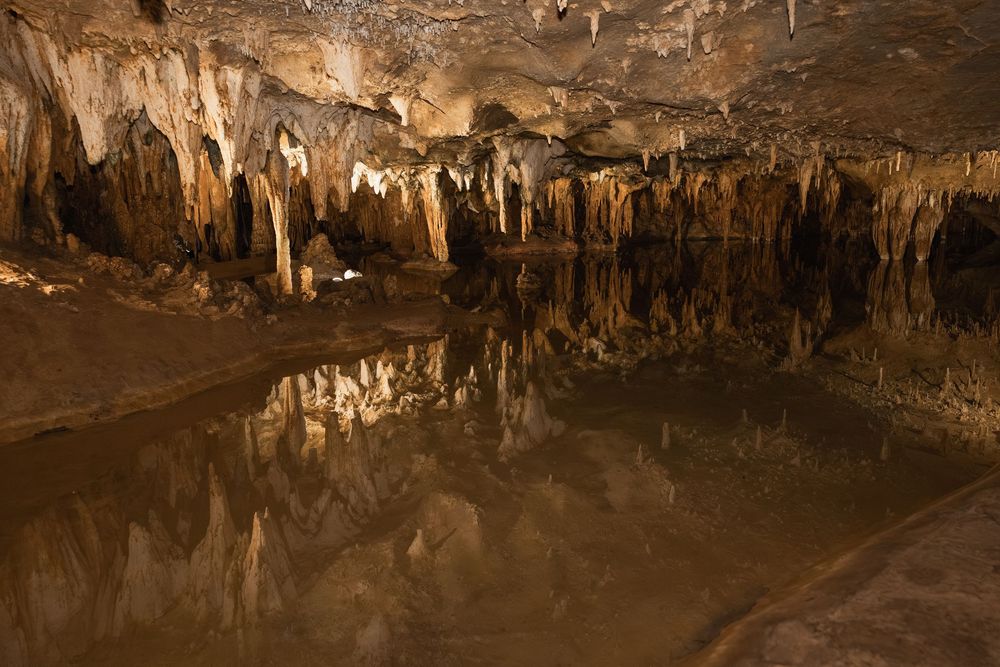 Dramatic limestone stalactites and stalagmites reflect in still cave waters creating a mirror-like underground landscape.