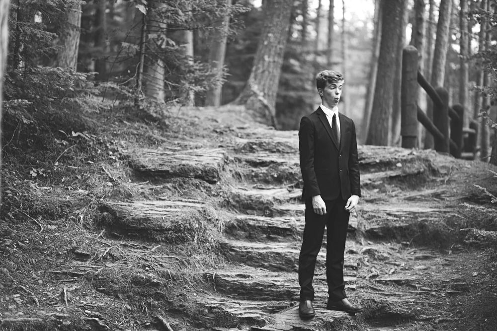 Black and white portrait of someone standing on a forest path among tall trees.