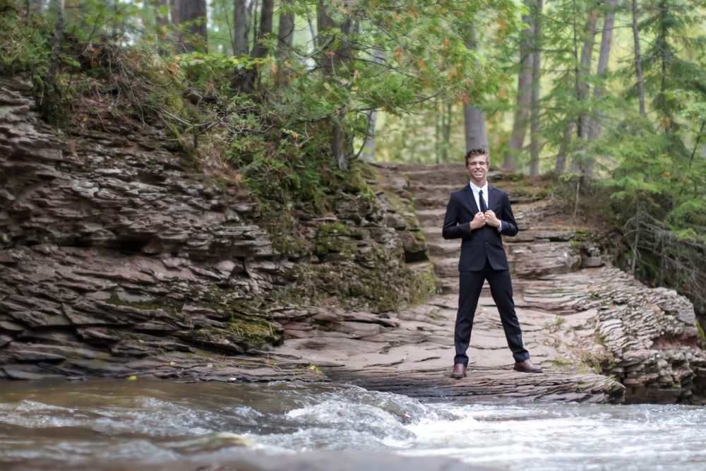 Person standing near rushing water and rocky cliffs in a forest setting.