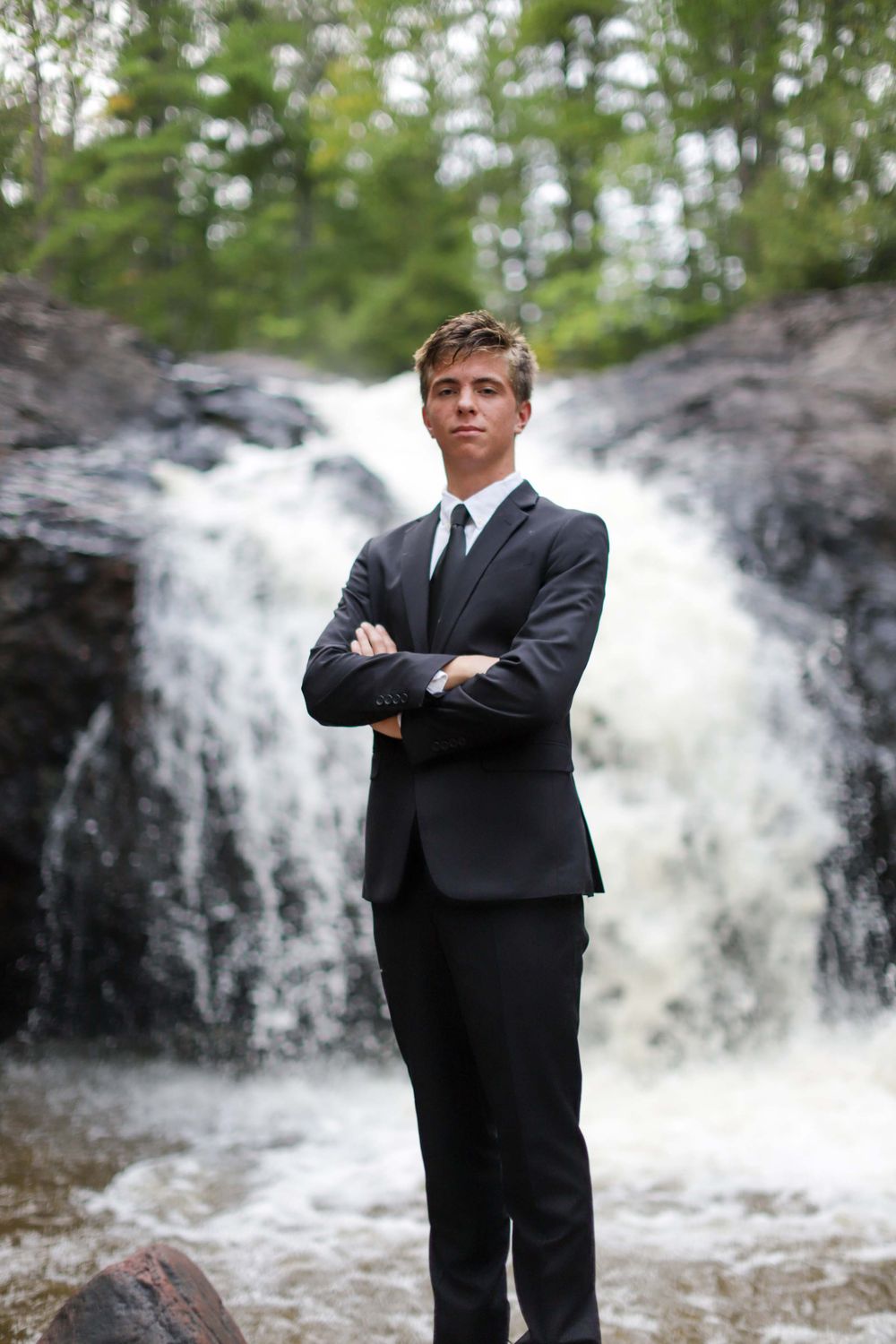 Figure in formal black suit poses in front of a rushing waterfall in natural surroundings.