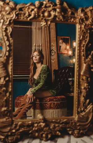 A person in a green velvet dress poses elegantly on a vintage couch surrounded by ornate gold-framed mirrors.
