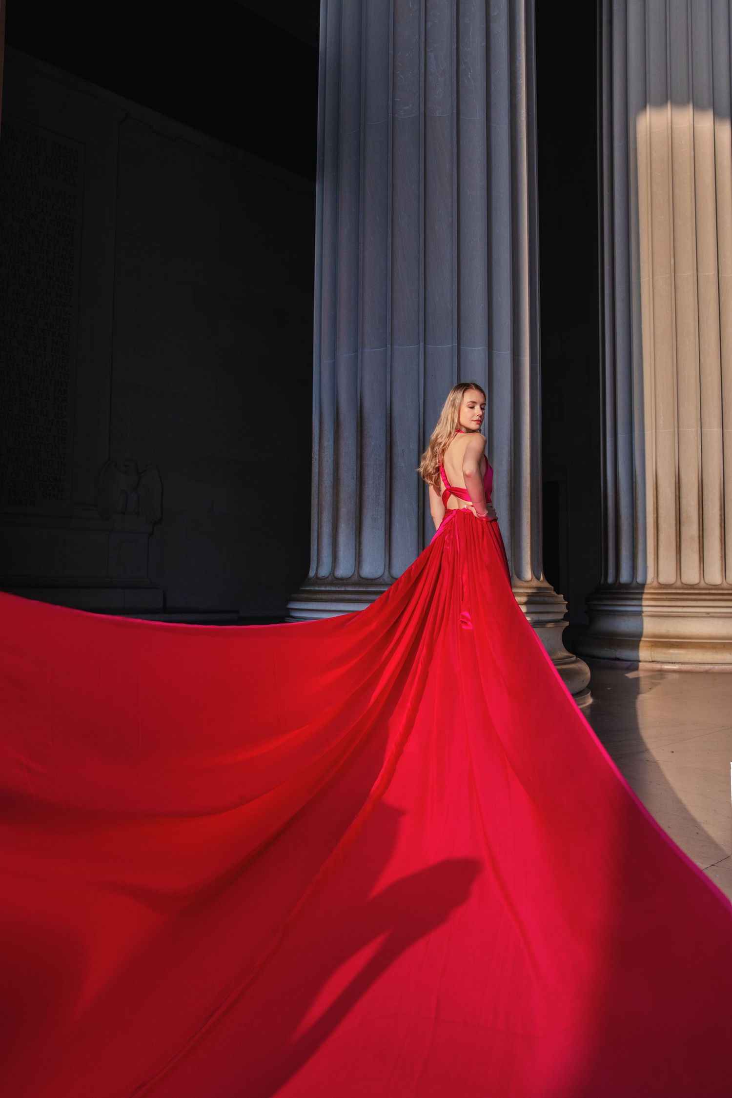 A flowing red gown with a dramatic train cascades across dark marble columns in an architectural setting. Flying Dress Photoshoot