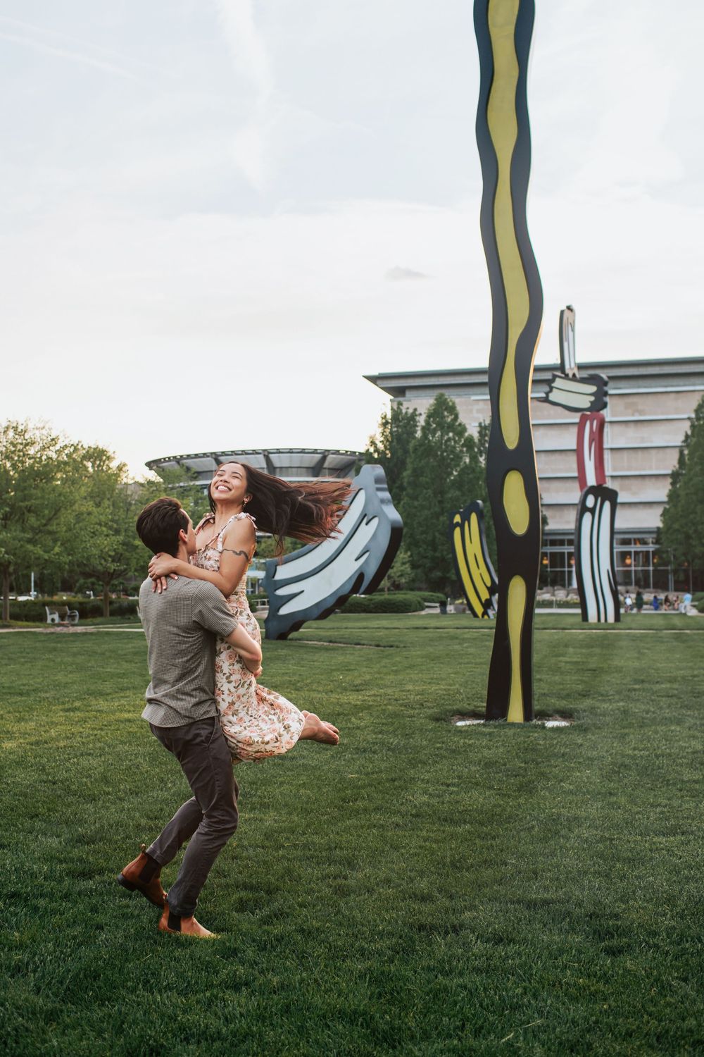 A couple dances playfully on a grassy lawn with large outdoor sculptures in the background.