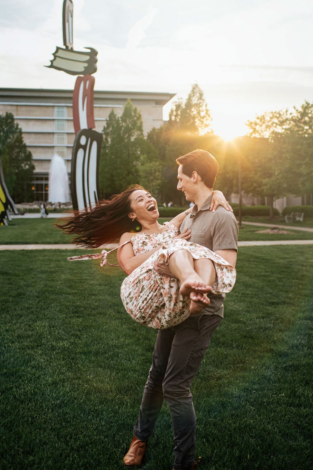 A couple dances playfully on a grassy lawn at sunset, with a building visible in the background.