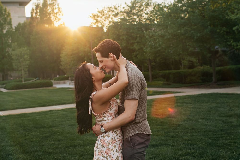 A couple embraces in a sunlit park during golden hour as the sun sets behind trees.