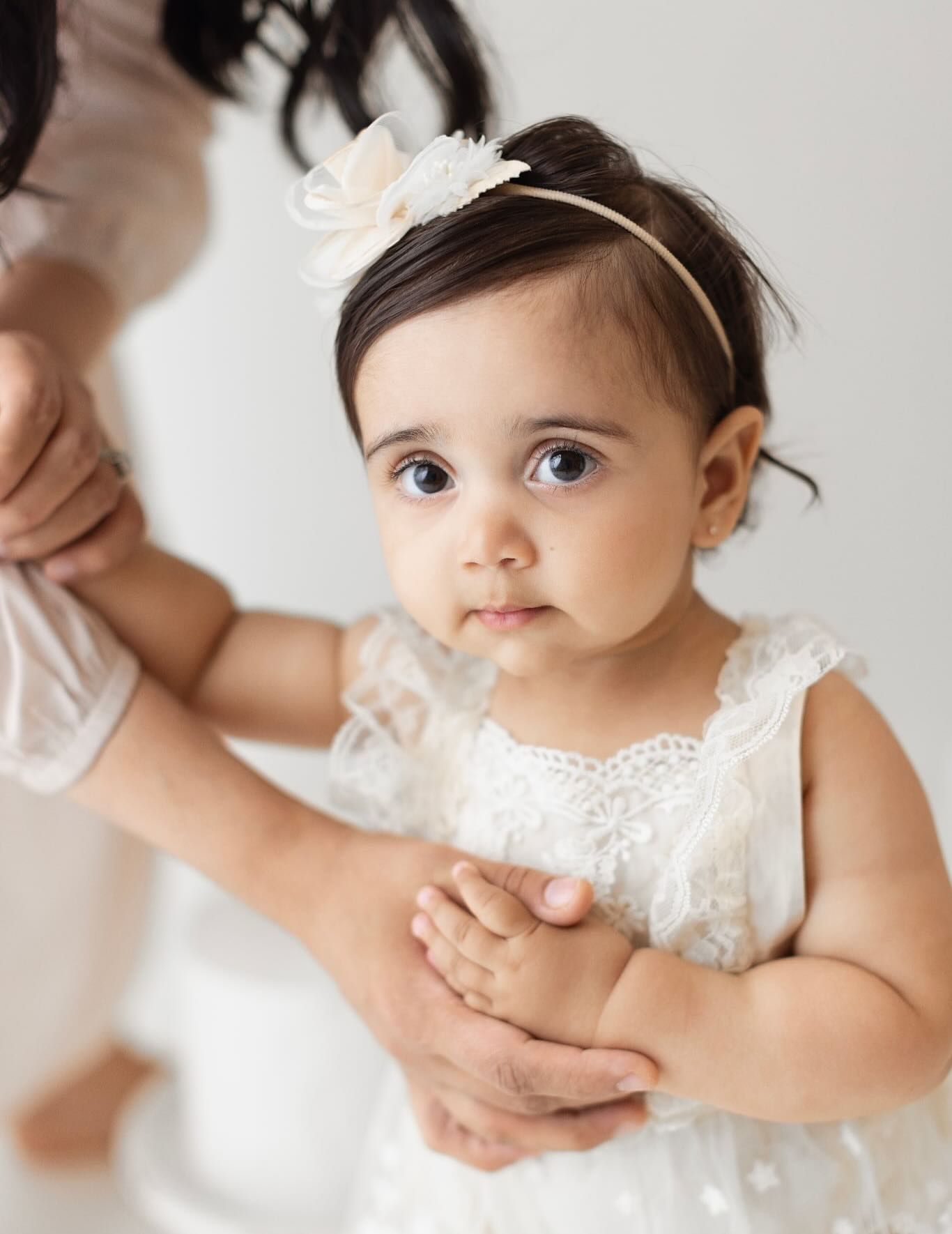 Baby in white lace dress and floral headband holds hands with someone while looking to the side.