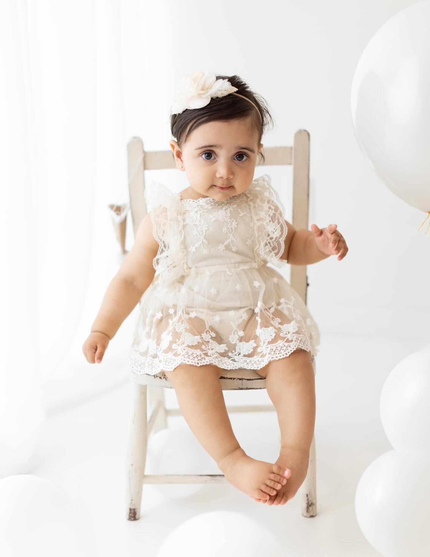 A baby wearing an elegant white lace dress sits on a wooden chair surrounded by white balloons.