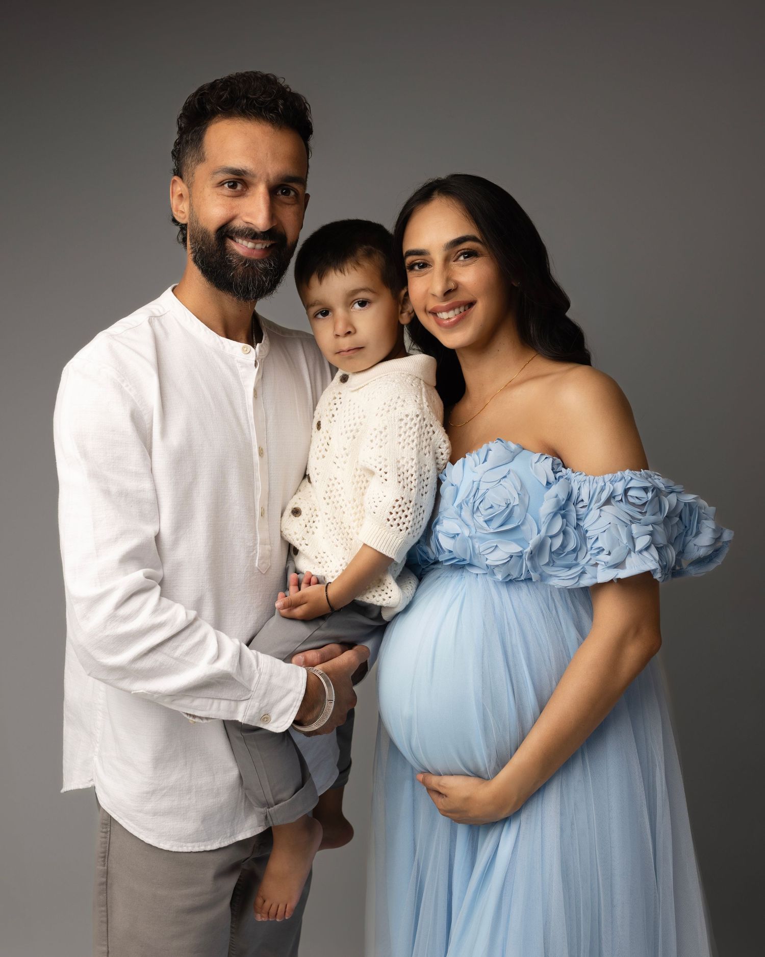 A family of three poses together in coordinated outfits of white and light blue against a neutral studio backdrop.