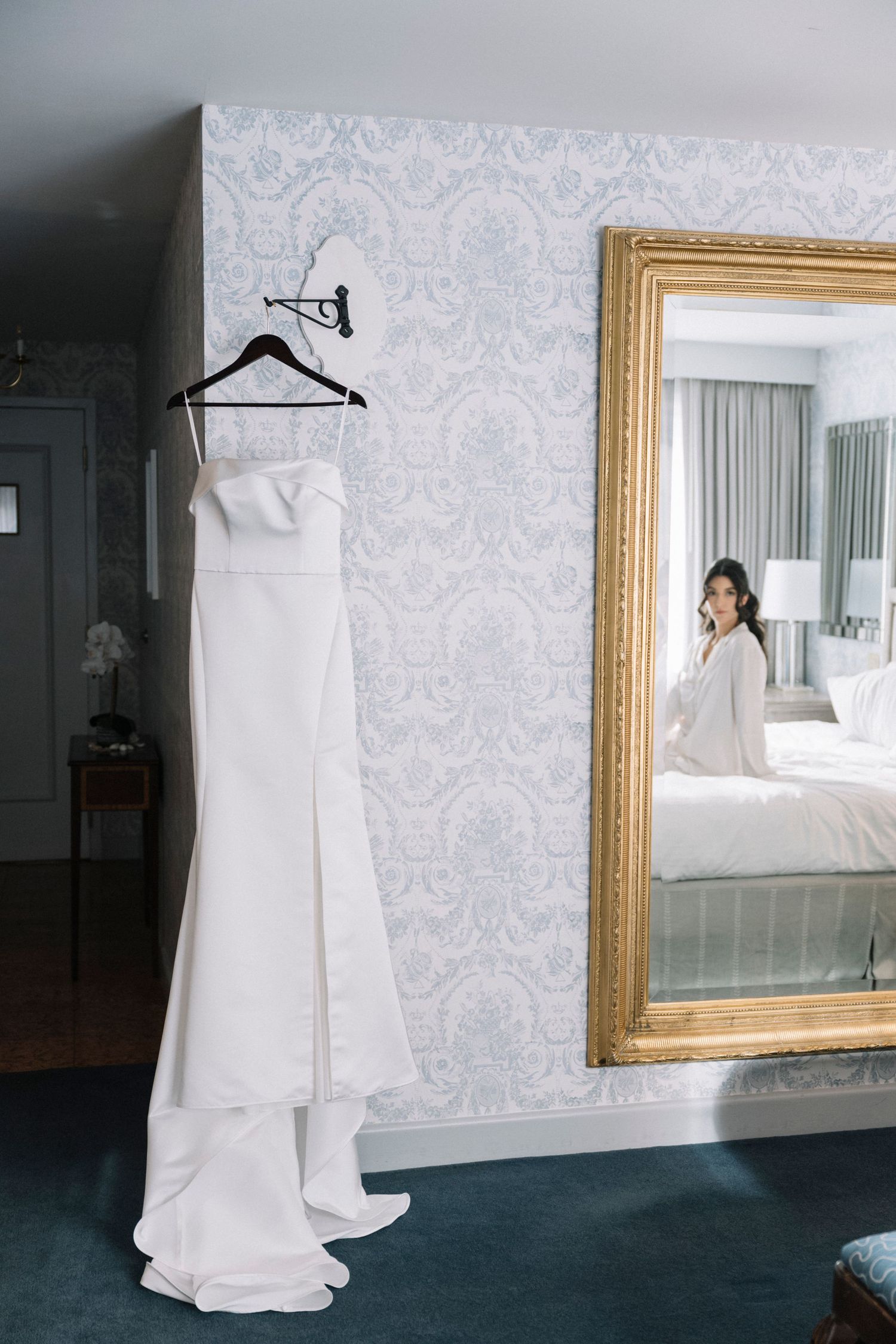 White wedding dress displayed against damask wallpaper next to a gold-framed mirror in a hotel room.