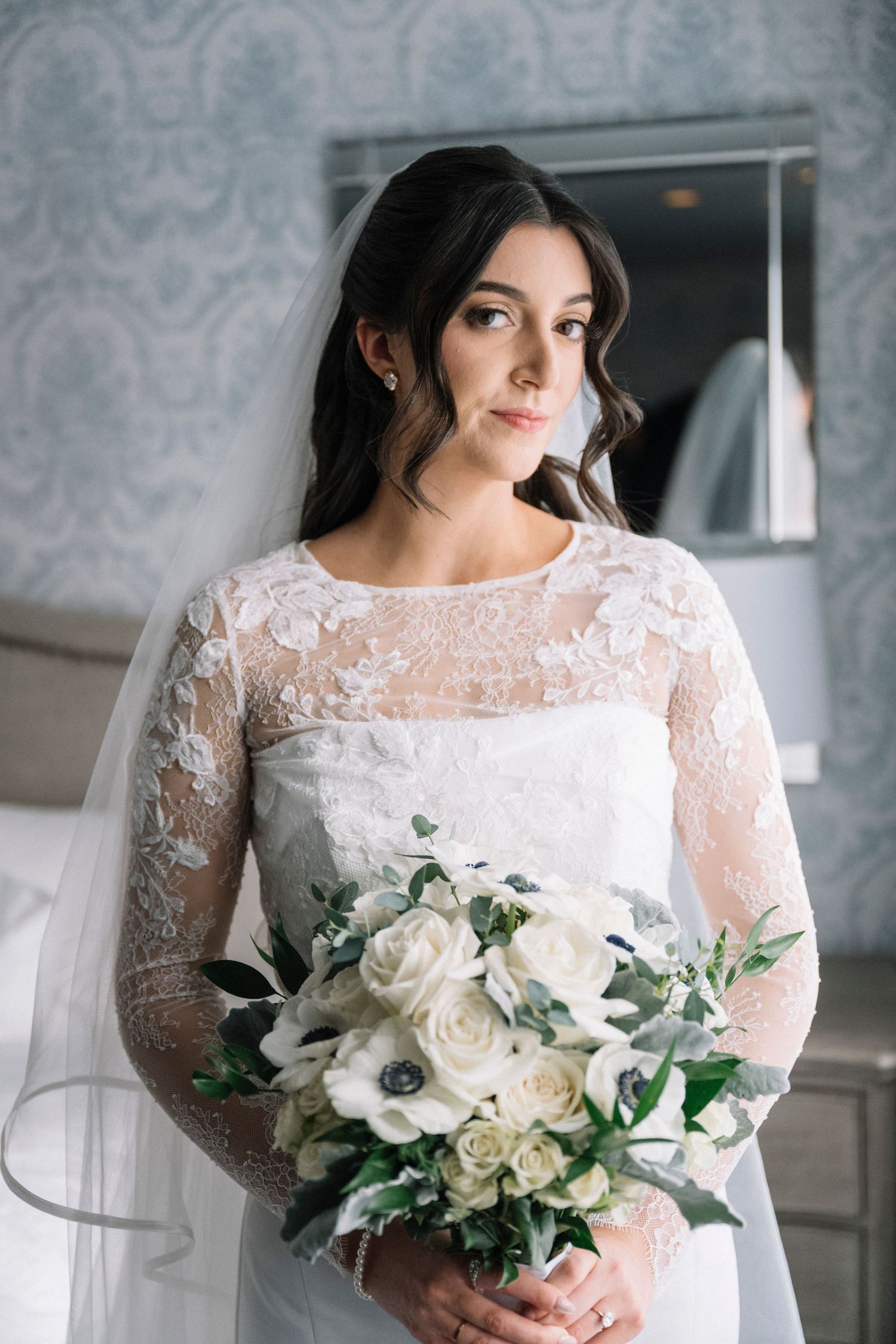 A bride in a long-sleeved lace wedding dress holds a luxurious white and green floral bouquet.