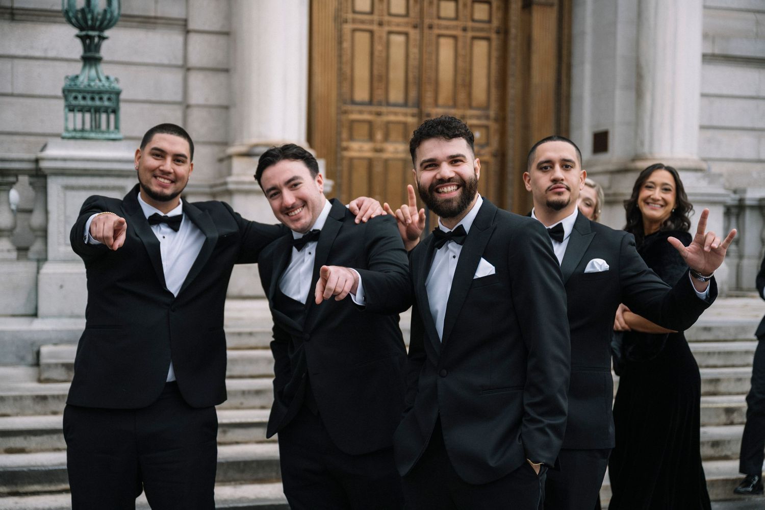 A wedding party in formal black suits and dresses poses together on stone steps outside an elegant building.